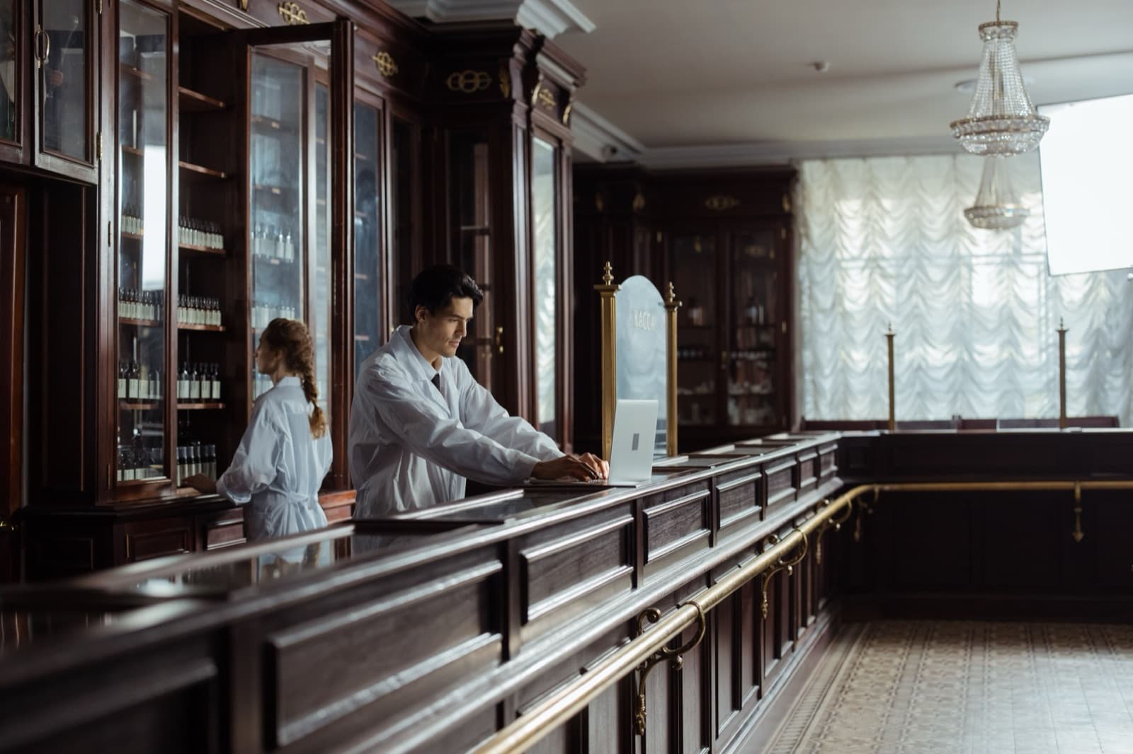 A pharmacist reviewing a prescription on a laptop at the pharmacy counter while a prior authorization request is processed